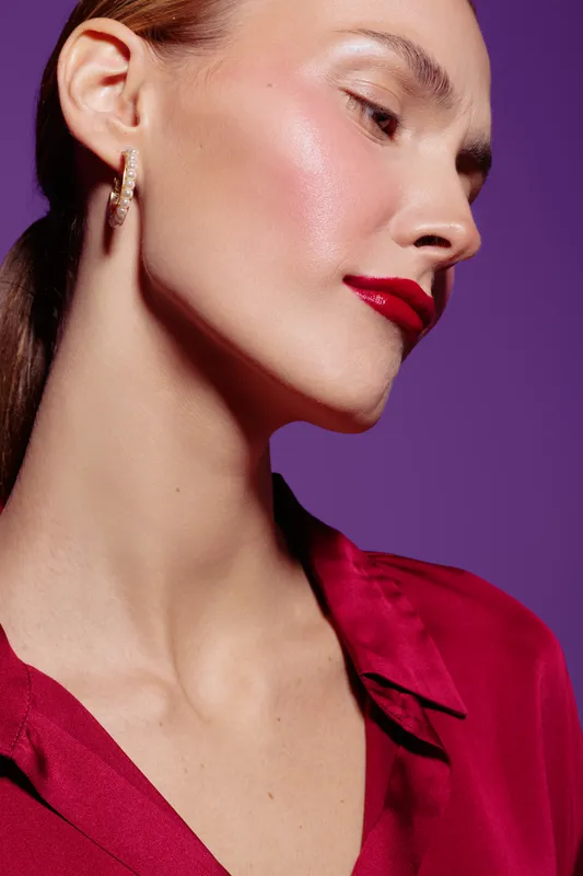 Close-up portrait of a woman with glossy skin and red lipstick wearing gold earrings and a red blouse against a purple background. Concept of beauty, fashion, and elegance.