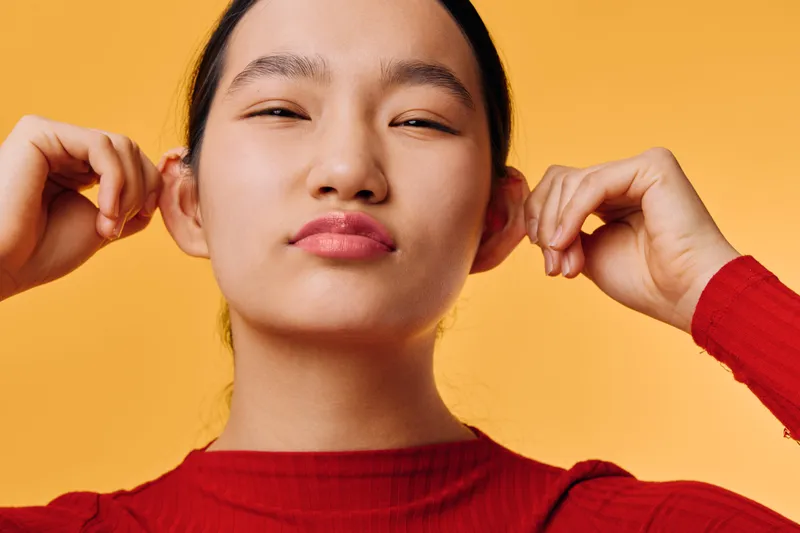 Young woman in red sweater with closed eyes making playful face and holding ears in studio with orange background. Portrait, fun, expression and casual fashion style with natural light.