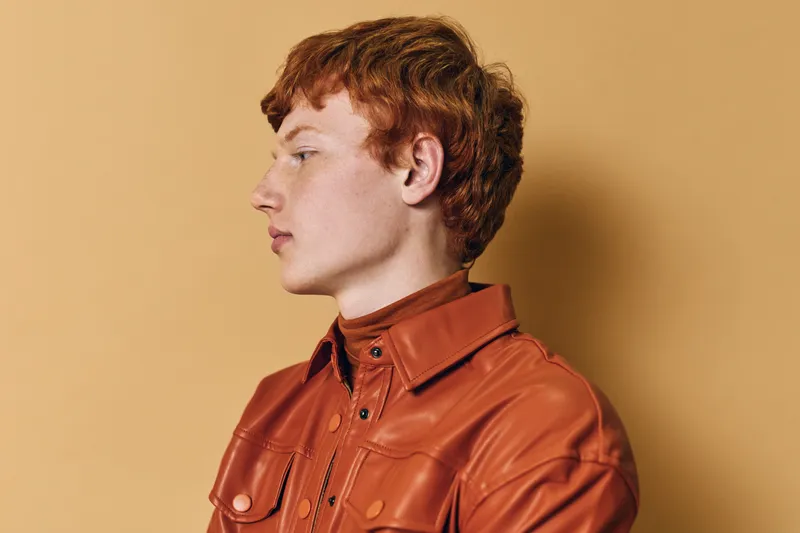 Portrait of young man with red hair wearing brown leather jacket standing against beige background with serious expression and side profile view in studio lighting.