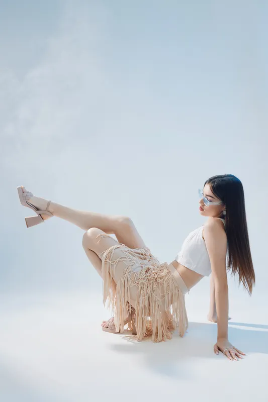 Young woman in white top and fringed skirt poses sitting with legs raised on light background. Concept of beauty fashion and modern style with sunglasses.