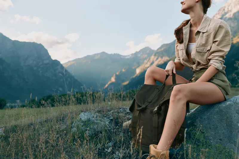 Young woman on a hike rests on a rock, wearing a casual jacket and shorts, backpack beside, overlooking rugged mountains and a grassy meadow in a peaceful outdoor scene.