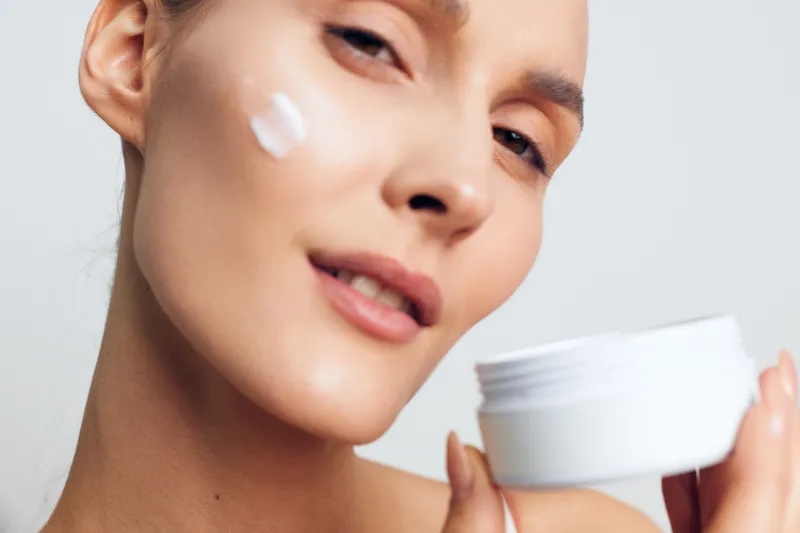 Young woman applies moisturizer cream on face holding white jar skincare beauty treatment isolated on light background with smooth healthy skin and natural makeup closeup portrait.
