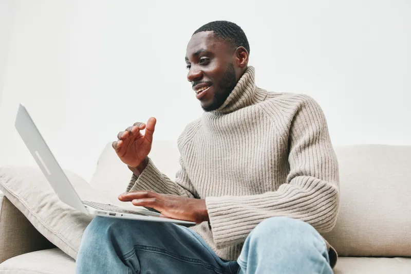 Young Black man in a cozy sweater smiles while using a laptop at home, showcasing a relaxed attitude and modern lifestyle