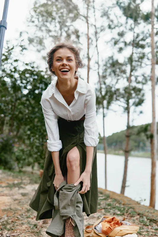 Happy woman in a green skirt and white shirt enjoying nature by the lake, joyful expression, outdoor setting, seasonal vibe, fashion details, lifestyle photography