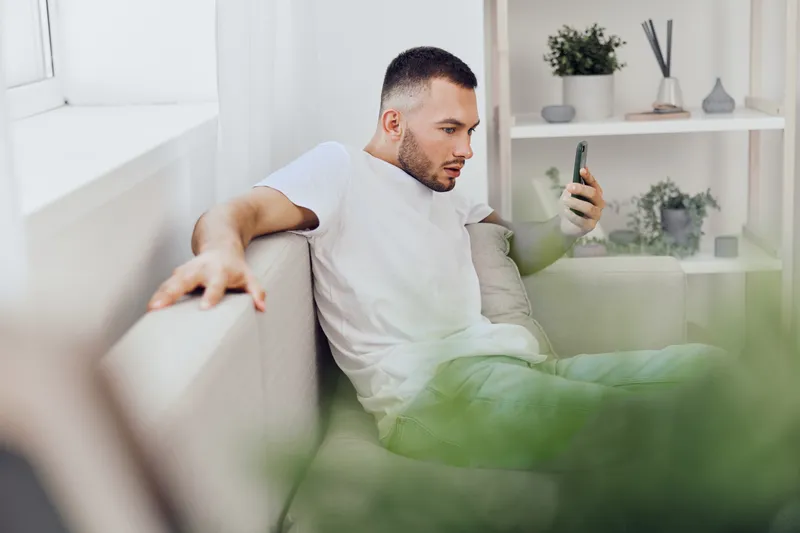 Focused man engaging with smartphone in a modern living room, surrounded by greenery. Perfect for concepts of technology and lifestyle in 2025-2026.