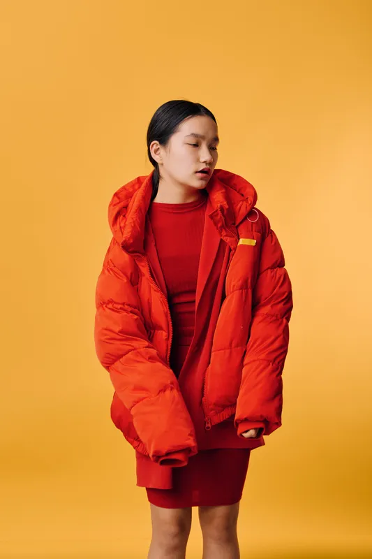 Young woman wearing oversized red puffer jacket and red dress posing against yellow background. Fashion model in warm outerwear with casual hairstyle looking aside for style photoshoot.