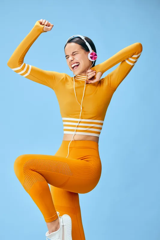 Energetic woman in bright orange fitness wear dances mid jump with headphones, capturing dynamic sport movement against a blue background for a lively workout lifestyle.