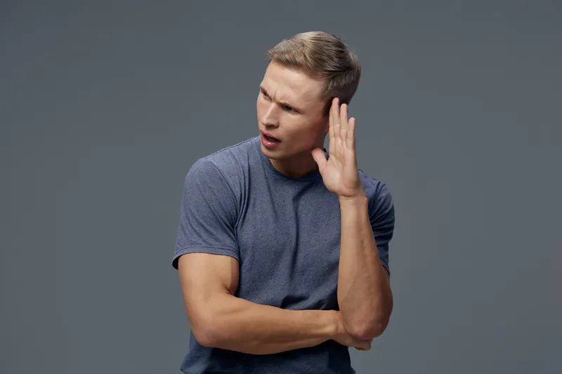 confused young caucasian man wear blue shirt listen hand near ear isolated on grey background studio portrait mental health concept