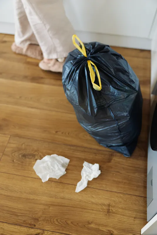 Garbage trash waste bag floor tissue near kitchen cabinet on hardwood, black tied plastic sack with yellow tie and crumpled paper on laminate, feet of person in background during home cleaning.