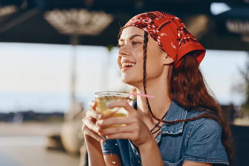 smiling woman holding a cold drink outdoors wearing a red bandana and denim shirt enjoying sunny weather at a summer cafe