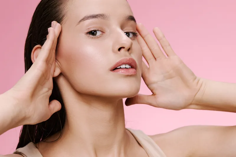 Beautiful young woman posing with hands on her face, showcasing a natural makeup look against a soft pink background, embodying beauty and self care