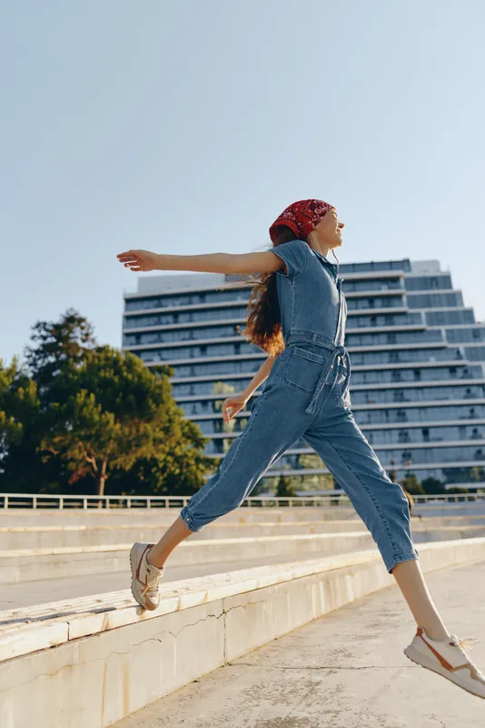 lifestyle woman in modern boho-western denim street style jumps with open arms outdoors, wearing a red bandana and sandals, film color, urban background.