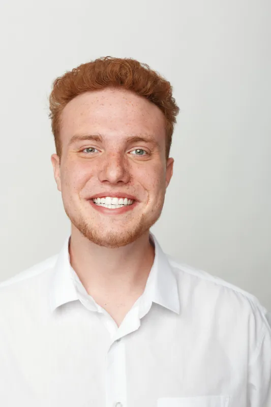 Smiling young man with red hair and beard wearing white shirt looks at camera with bright expression. Studio portrait on light background. Concept of happiness, confidence, and casual style.