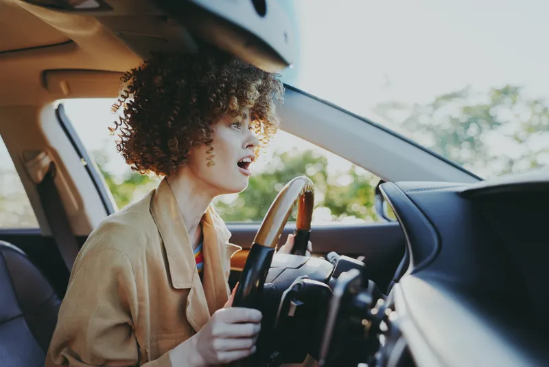 Woman driving a car, candid lifestyle moment with retro film color, warm tones on sunlit dashboard, relaxed expression, casual jacket, road ahead, urban vibe