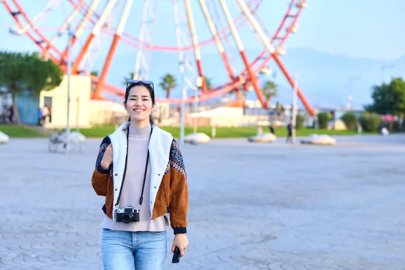 Woman with camera outdoors in a park with a large ferris wheel, casual jacket and jeans, smiling while strolling along a sunny plaza, travel vibe, candid lifestyle photography for leisure and
