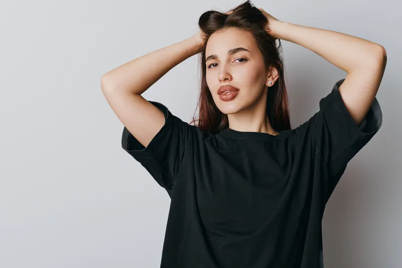 Young woman with brown hair posing confidently with hands behind head wearing black t shirt against plain light background. Female model with natural makeup and relaxed expression.