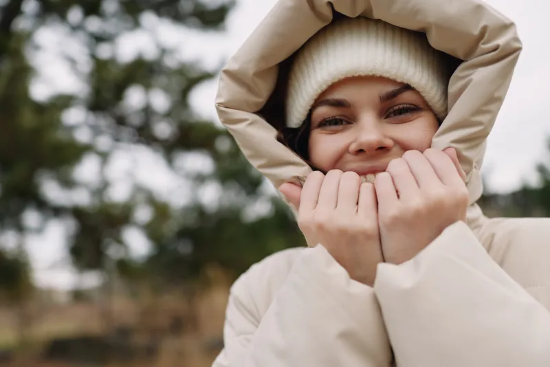 Winter coat hooded smiling outdoor portrait of a young woman in a warm jacket with a beanie embracing the chilly air close crop outdoors with blurred trees and a cozy casual mood