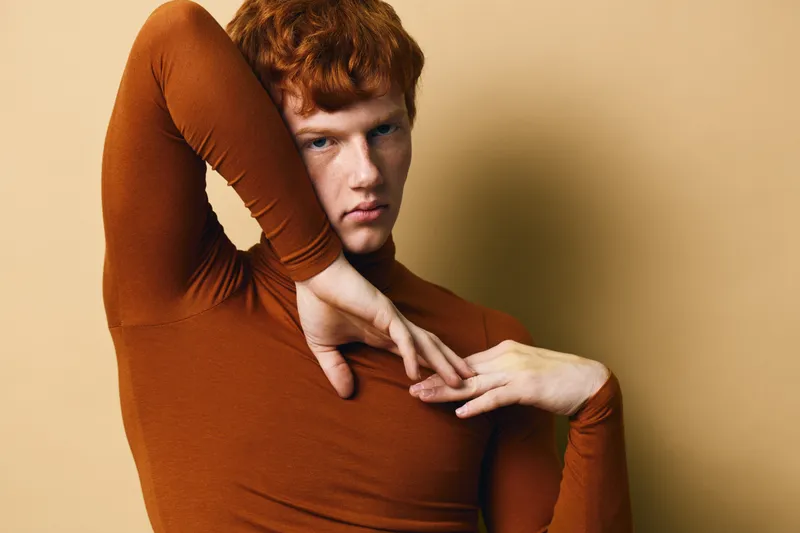 Redhead man with curly hair wearing brown turtleneck posing with hands near face against beige background. Fashion, portrait and studio photo of confident male model with intense look and stylish