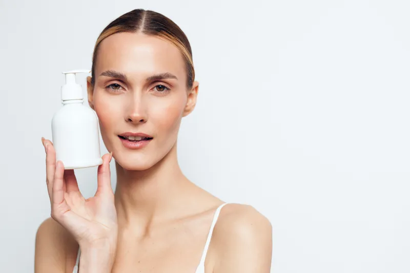 Young woman with clear skin holding white cosmetic bottle near face, natural makeup, smooth complexion, minimalistic background, skincare routine, beauty product concept