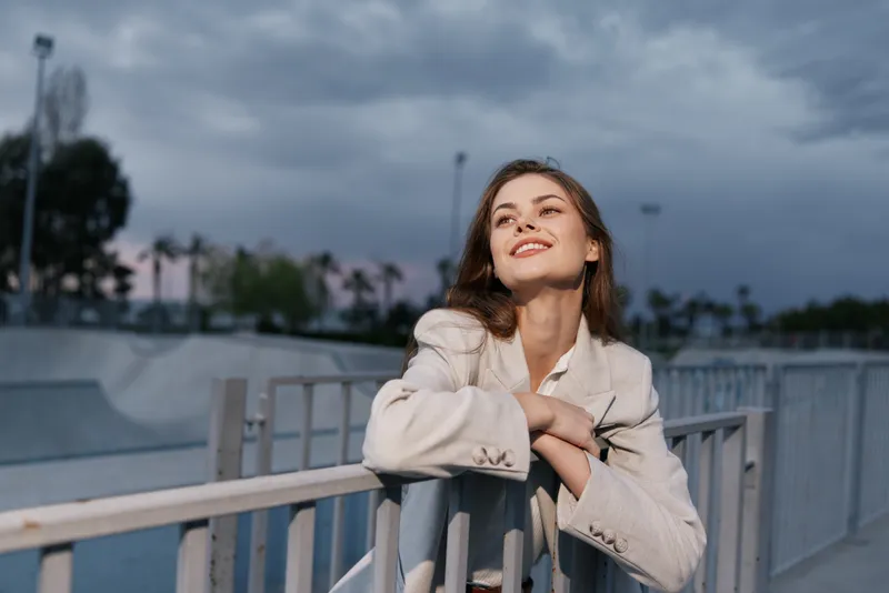 Young smiling woman in light blazer outdoors at dusk with thoughtful confident expression and urban blurred background, natural lifestyle portrait