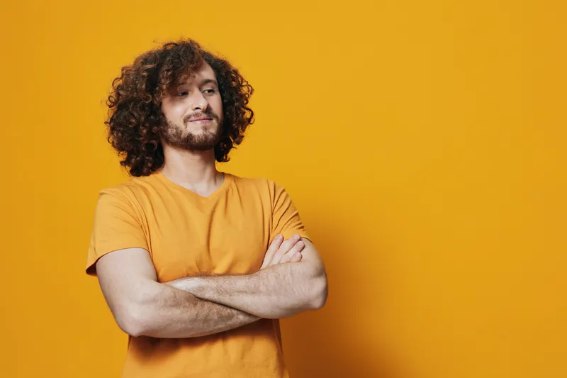 Confident young man with curly hair wearing a bright orange shirt against a vibrant yellow background, showcasing a positive emotion and seasonal style