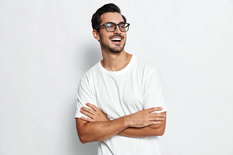 Smiling young man with stylish glasses poses confidently, arms crossed, wearing a casual white t-shirt against a minimalistic light background. People lifestyle concept.
