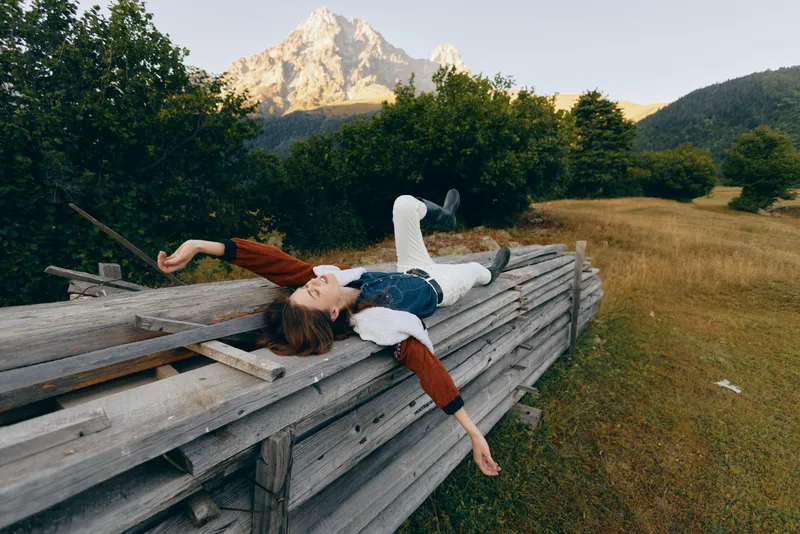Woman lying relaxed on wooden fence in meadow with mountain backdrop, portrait of outdoors leisure and nature escape, casual clothing and serene summer landscape.