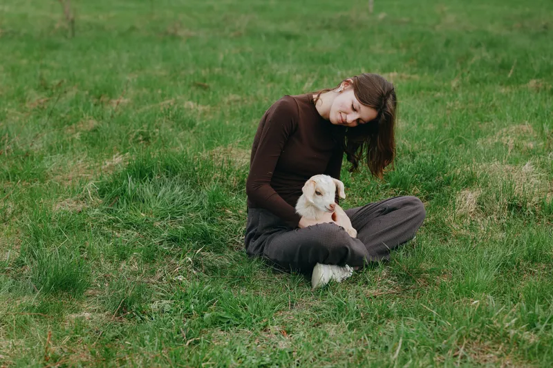 Joyful woman outdoors with a gentle goat kid, seated on sunlit grass, relaxed posture, warm tones, soft breeze, wholesome countryside moment, natural connection