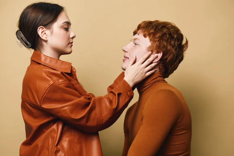 Young woman in orange leather jacket gently holds face of red-haired man in brown turtleneck during intimate moment with plain beige background in studio.