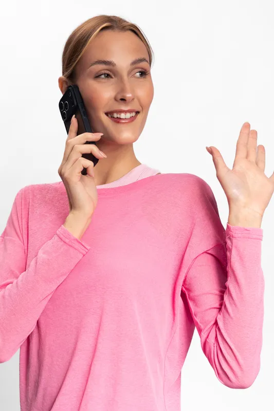 Young woman in pink sweater talks on phone and waves hand with smile. Isolated on white background. Concept of communication, happiness, casual style, and friendly gesture.