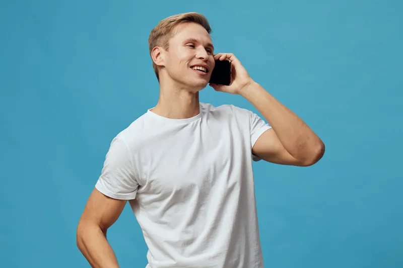 Smiling young man with short blond hair wearing casual white t-shirt talking on a mobile phone while standing against a bright blue background. Studio portrait showcasing casual communication and posi