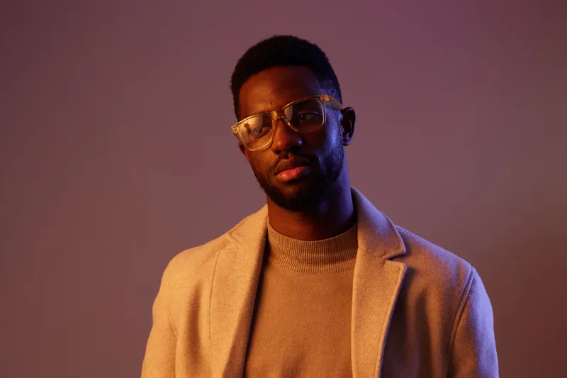 Stylish black man in glasses wearing a beige sweater and coat, posing with a serious expression against a purple background