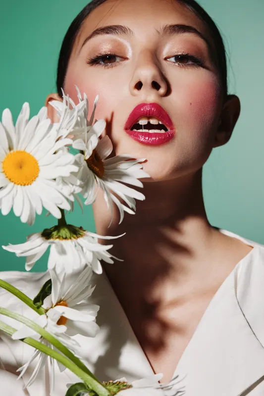 Close-up portrait of a young woman with glossy red lips and shimmering eyeshadow holding white daisies. Studio fashion shot on pastel green background capturing beauty creativity.