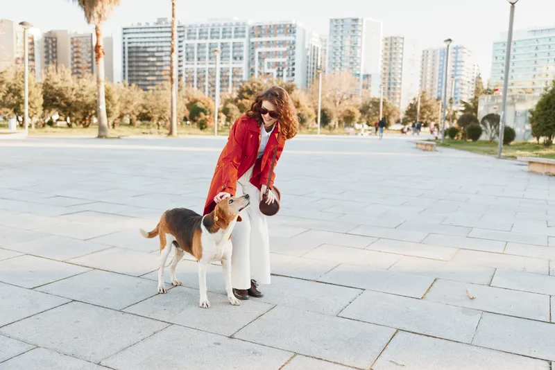 Young woman with curly hair wearing a red coat plays with her dog on a city square under bright daylight, showing joy and connection with her pet.