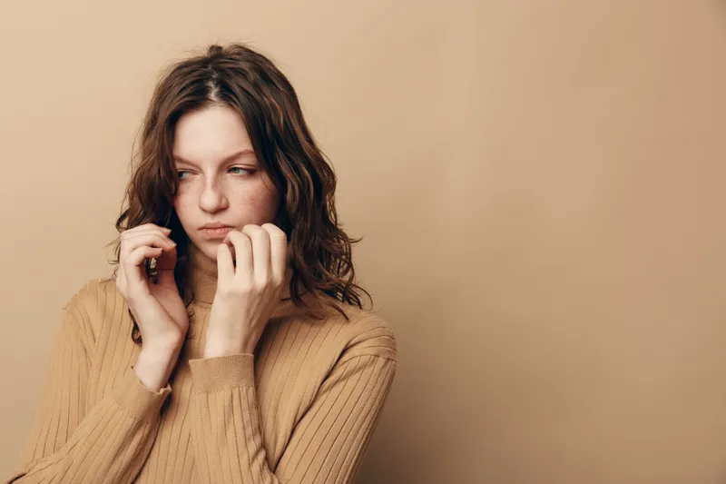 Sad young woman with wavy hair wearing beige sweater looks away thoughtfully on beige background. Concept of loneliness, contemplation, and emotional struggle in casual style.