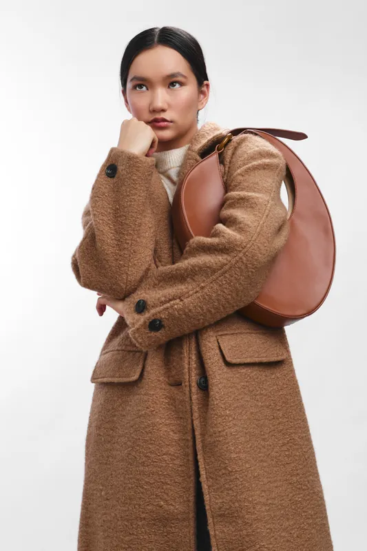 asian woman wearing a long tan coat with a round leather bag over her shoulder stands in a clean studio, presenting a poised, stylish and contemporary fashion moment.