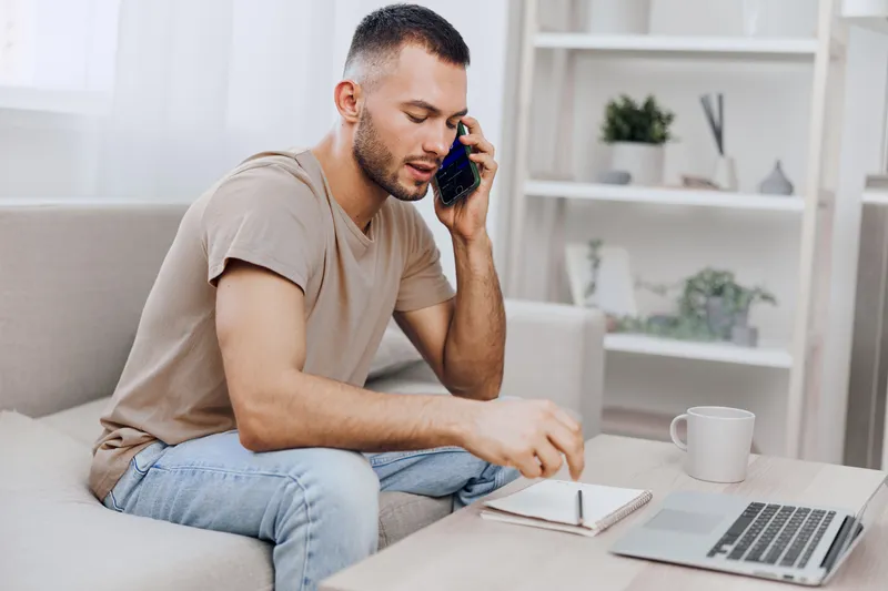 A focused man engaged in a phone conversation, making notes on a notebook while sitting comfortably on a sofa. Ideal for remote work or modern lifestyle themes.