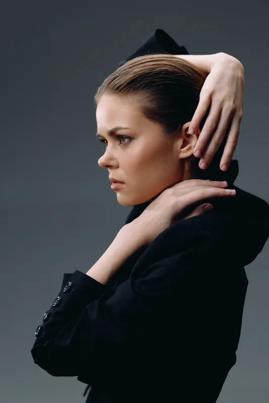 Profile of a serious young woman with slicked-back hair wearing a black blazer against a dark background.