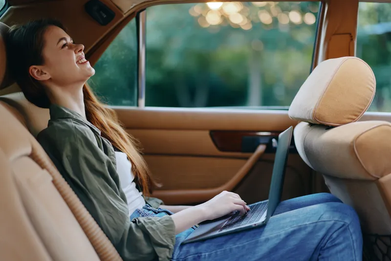 Young woman working on a laptop inside a car, smiling with relaxed expression, enjoying the moment outdoors, capturing the essence of freedom and creativity.