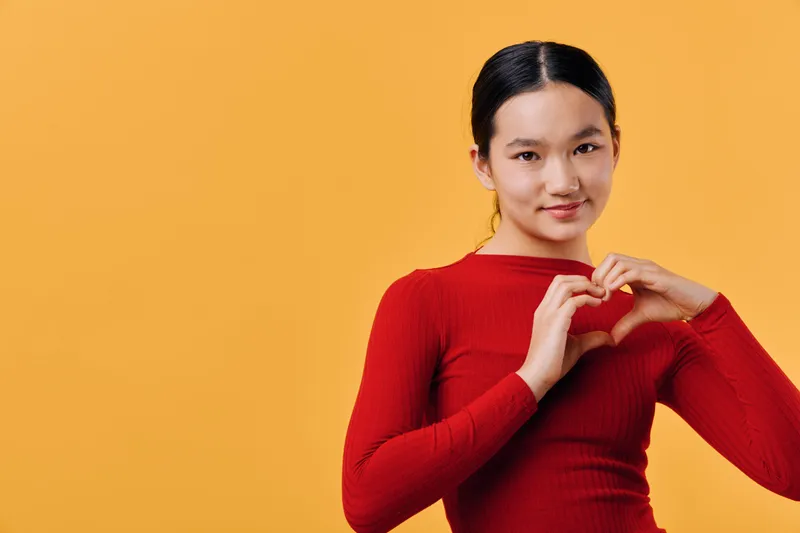 Young woman in red sweater making heart shape with hands against orange background, smiling and looking at camera with positive expression and casual hairstyle.