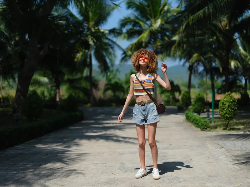 young woman with curly hair wearing colorful striped crop top and denim shorts walking outdoors on sunny day, smiling and enjoying warm weather, tropical palm trees background