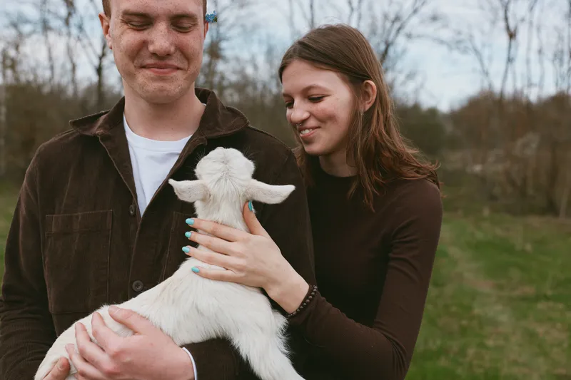 Goat kid with a content couple outdoors in a sunlit field, a gentle breeze and warm smiles. A candid lifestyle moment showing tenderness, affection, and natural interaction with animals.