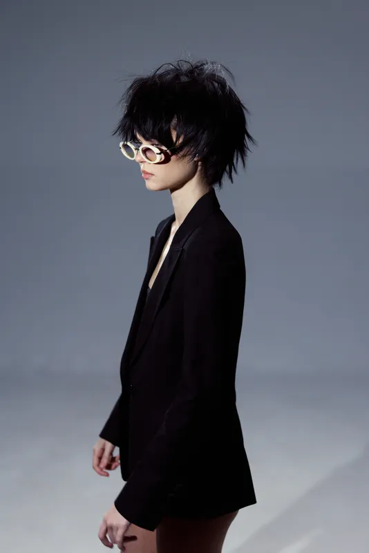 Stylish woman with short messy hair wearing oversized glasses and a black blazer in a minimalistic setting