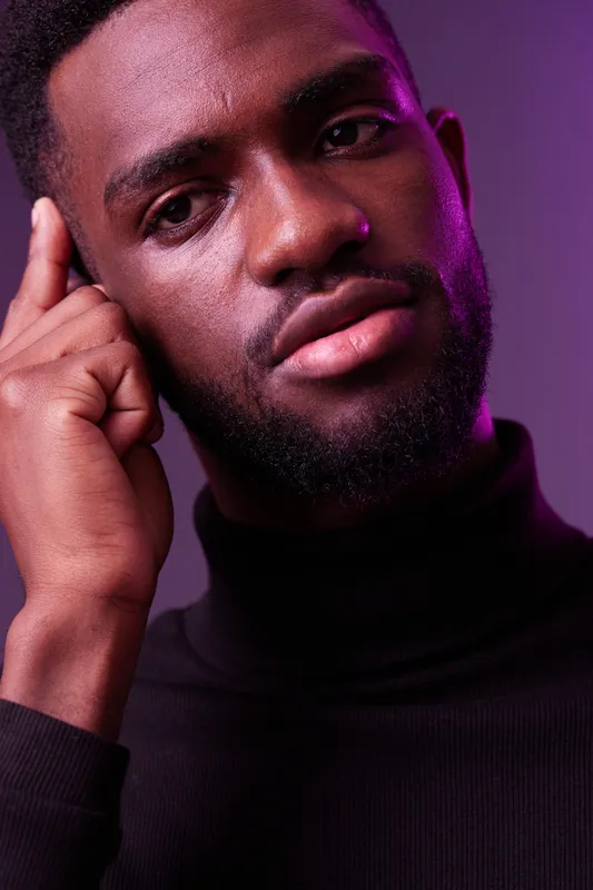 A thoughtful young Black man in a black turtleneck gazing directly, set against a vibrant purple backdrop, conveying introspection and creativity