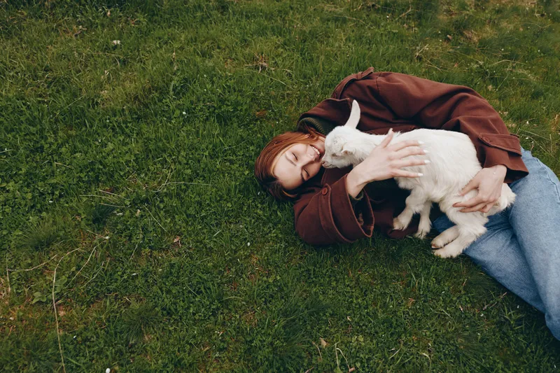 A woman lies on the grass, hugging a white goat kid with gentle affection. Outdoor scene portraying calm lifestyle, nature, and animal companionship under soft daylight.