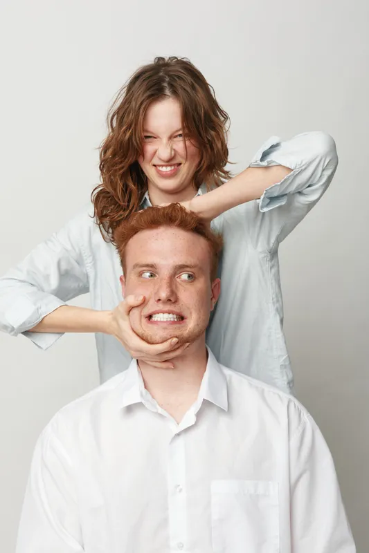 Playful young woman with curly hair squeezes redhead man face with hands. Both wear white shirts and pose on light background. Concept of friendship, fun, humor, and casual lifestyle.