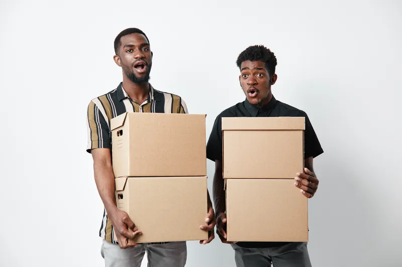 Two young black men with dark skin holding cardboard boxes for moving or delivery on white background. African ethnicity, casual clothing, teamwork and logistics concept.