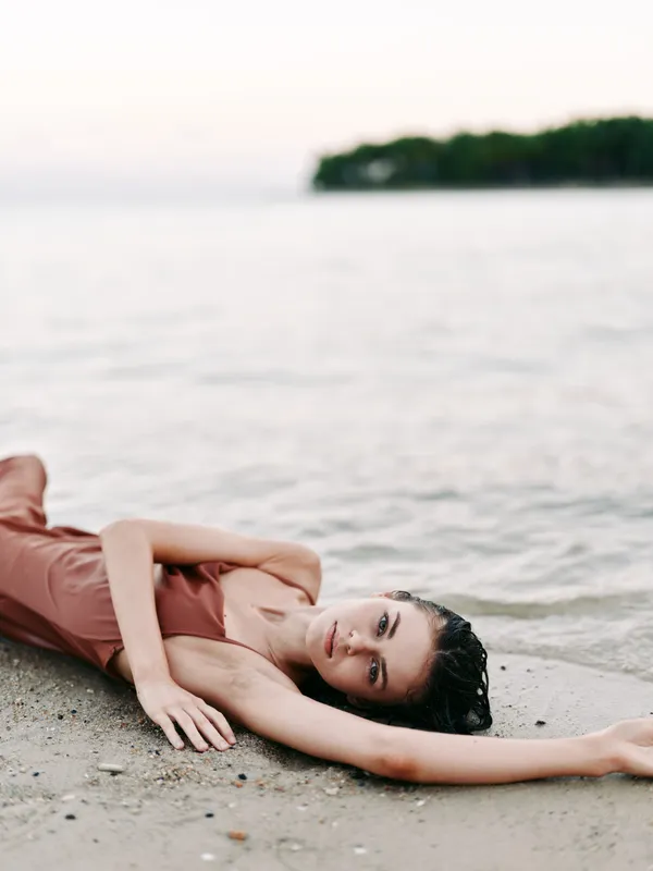 A relaxed woman lies on a sandy beach by the calm shoreline, wearing a soft dress, arms extended, hair wet, sunlight and sea creating a serene, peaceful scene.