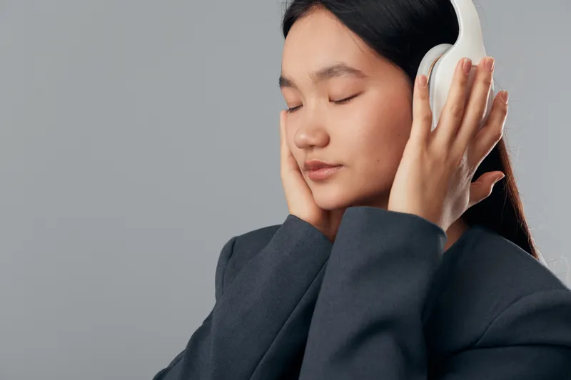 Woman wearing headphones with closed eyes enjoying music or meditation in casual dark jacket against plain gray background with peaceful expression and hands adjusting ear cups.