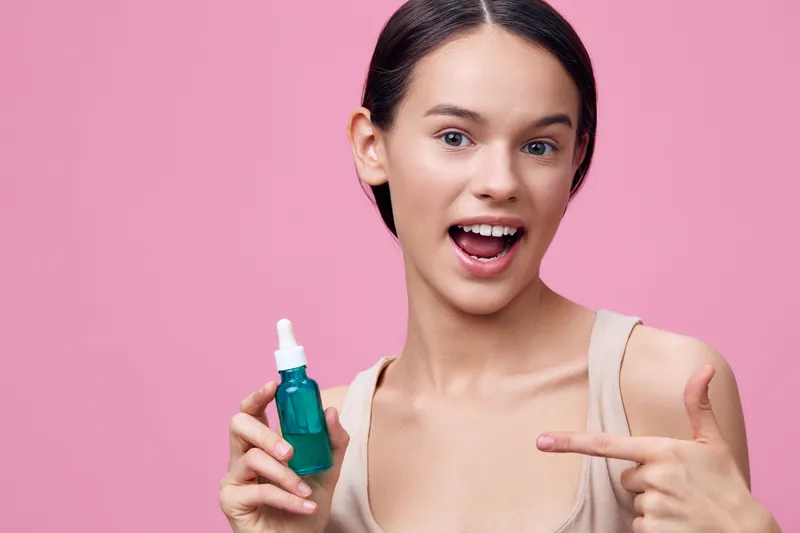 Young woman promoting skincare serum with a vibrant smile and pointing, against a pink background, showcasing beauty, skincare, and healthy skin